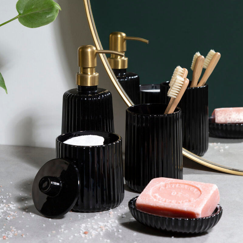 Black bathroom accessories including bottles, a soap dish, and toothbrush holder on a gray surface with a plant in the background.