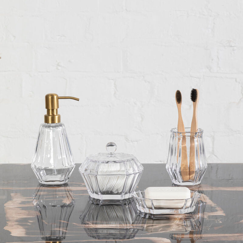 Bathroom accessories including a soap dispenser, glass containers, and toothbrushes on a marble surface with a white brick wall background.