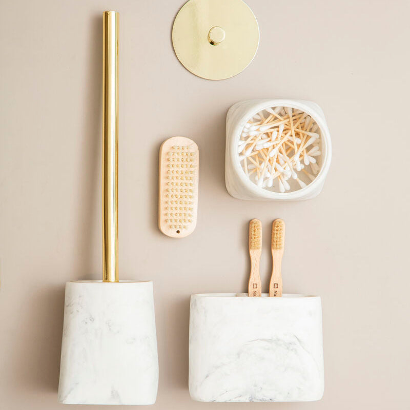 Bathroom accessories including a marble holder with a gold-plated brush, a small round holder with a candle, a white container with wooden toothbrushes, and a wooden scrubber on a beige background.