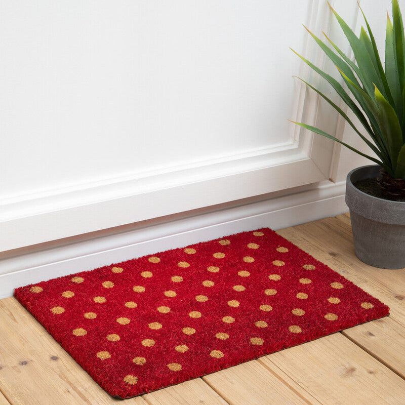 Red doormat with gold polka dots on a wooden floor next to a plant.