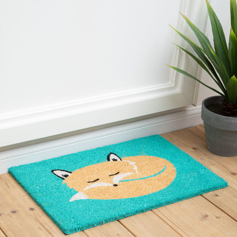 Teal doormat with fox design on a wooden floor next to a plant.