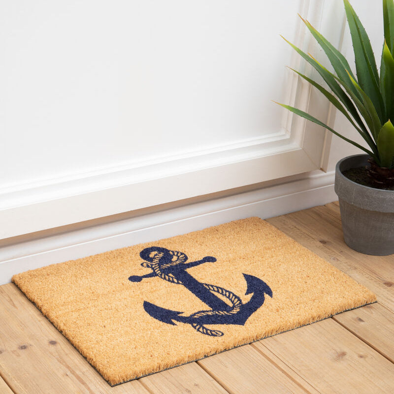 Doormat with an anchor design on a wooden floor next to a plant.