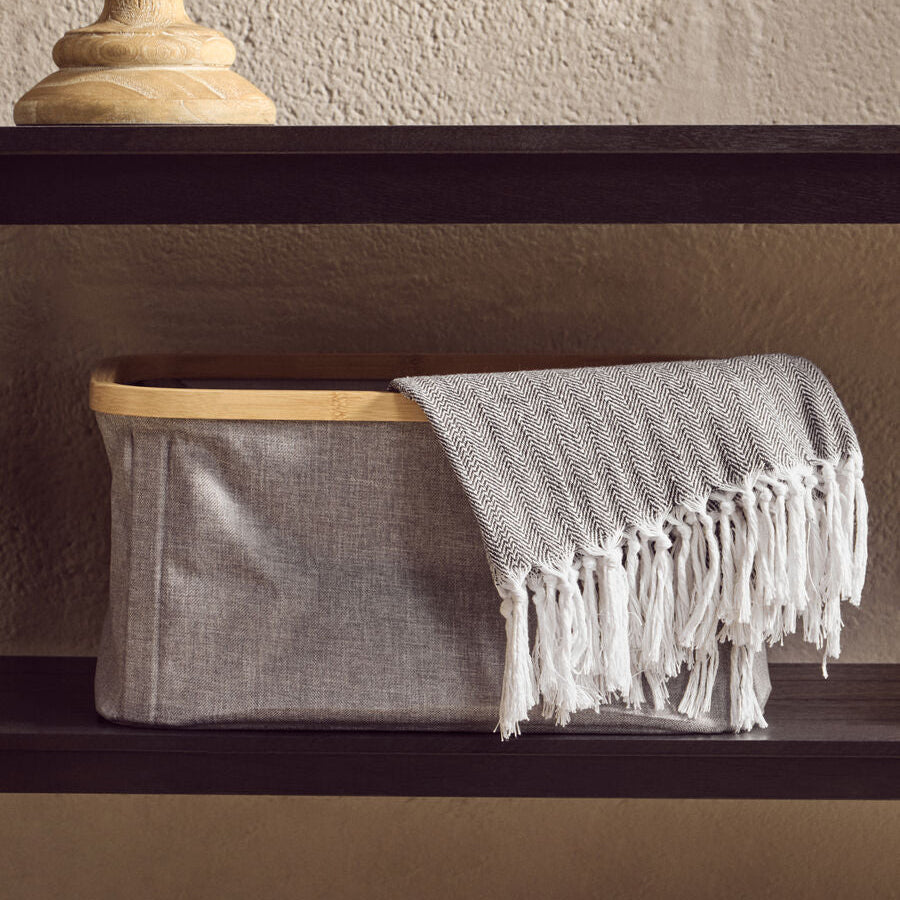 Gray fabric basket with a white fringed towel on a dark shelf against a beige wall.