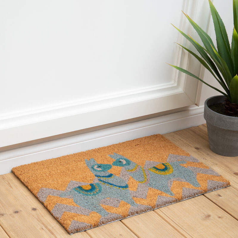 Decorative doormat with colorful pattern on a wooden floor next to a plant.