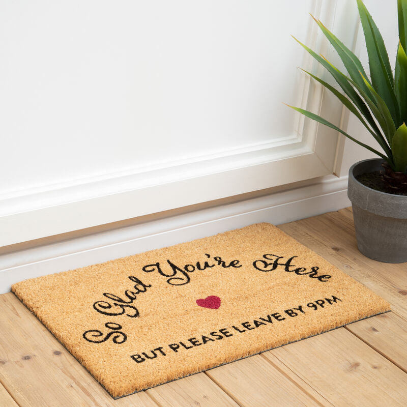 Doormat with humorous text on a wooden floor next to a plant