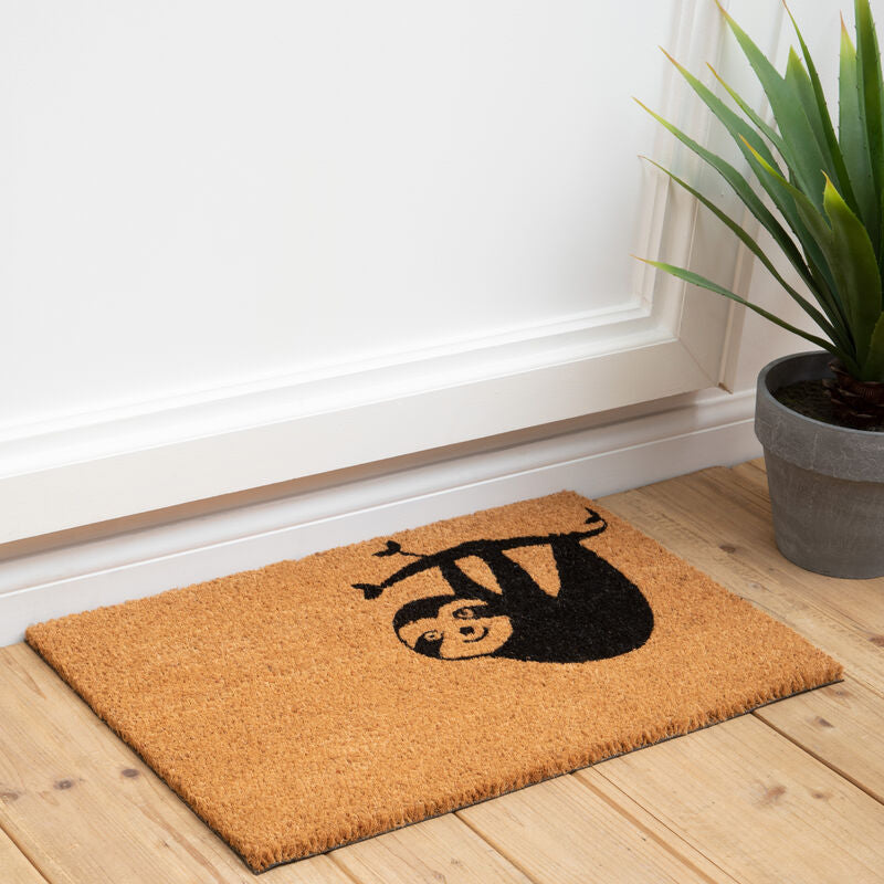 Doormat with a black design on a wooden floor next to a plant
