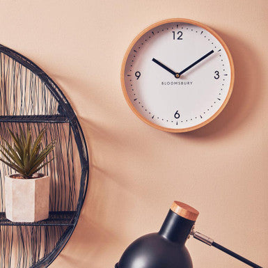 Wall clock with wooden frame and black hands on a beige wall, next to a black lamp and plant.