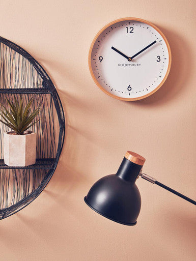 Wall clock with wooden frame and black hands on a beige wall, next to a black lamp and plant.