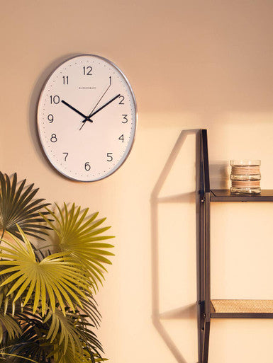 Wall clock on a beige wall with a plant and shelf in the foreground