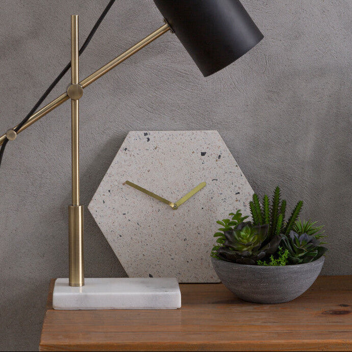 Table lamp with black shade and gold arm on a wooden table against a gray wall.