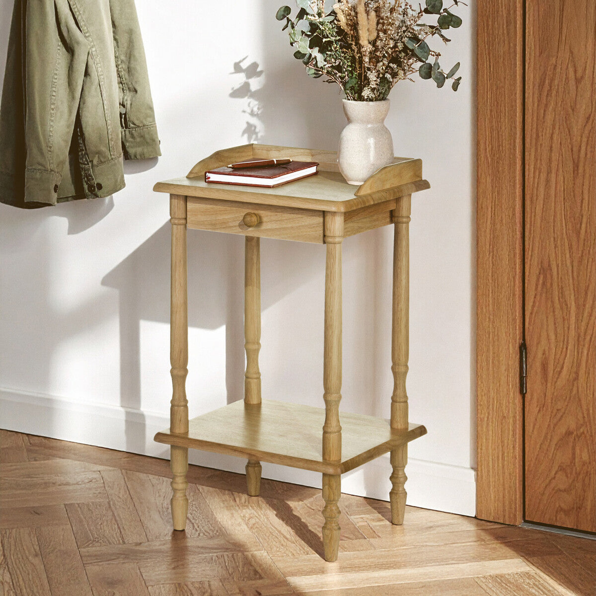 Wooden side table with a vase of flowers and a book in a room with wooden flooring and a white wall.