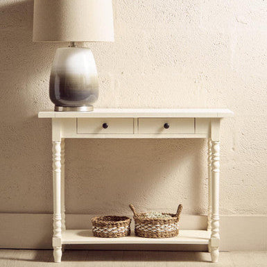 White console table with a lamp and two baskets against a beige wall.