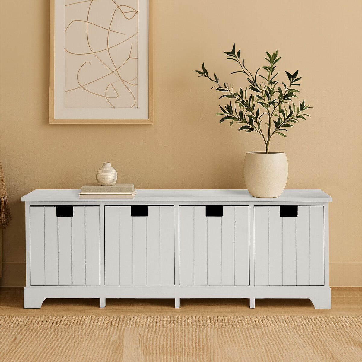 White storage bench with black handles against a beige wall with abstract art and a plant.