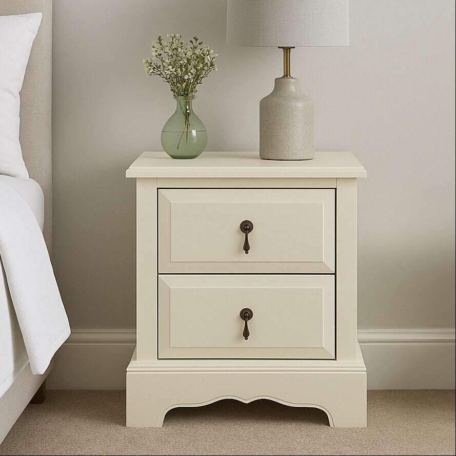 White nightstand with a lamp and vase next to a bed in a bedroom setting.