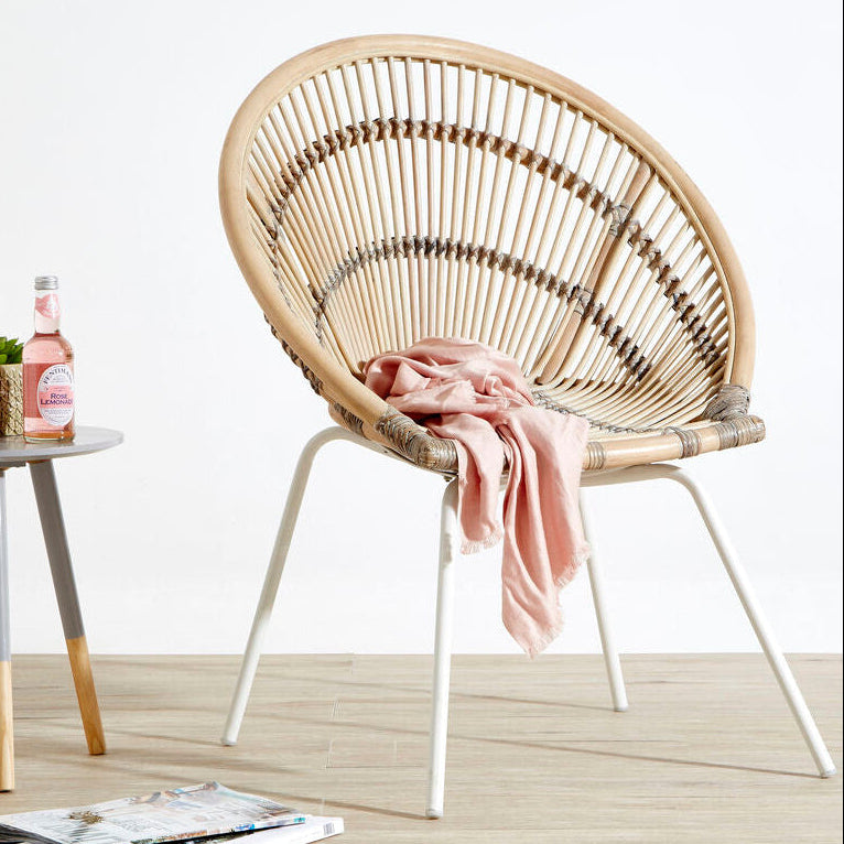 Wicker chair with a pink blanket on a wooden floor, next to a small table with a bottle and plant.