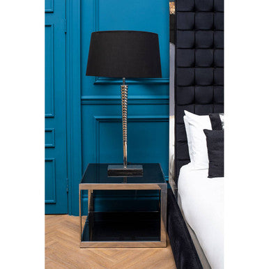 Black table lamp on a glass nightstand against a blue wall with a bed in the background.