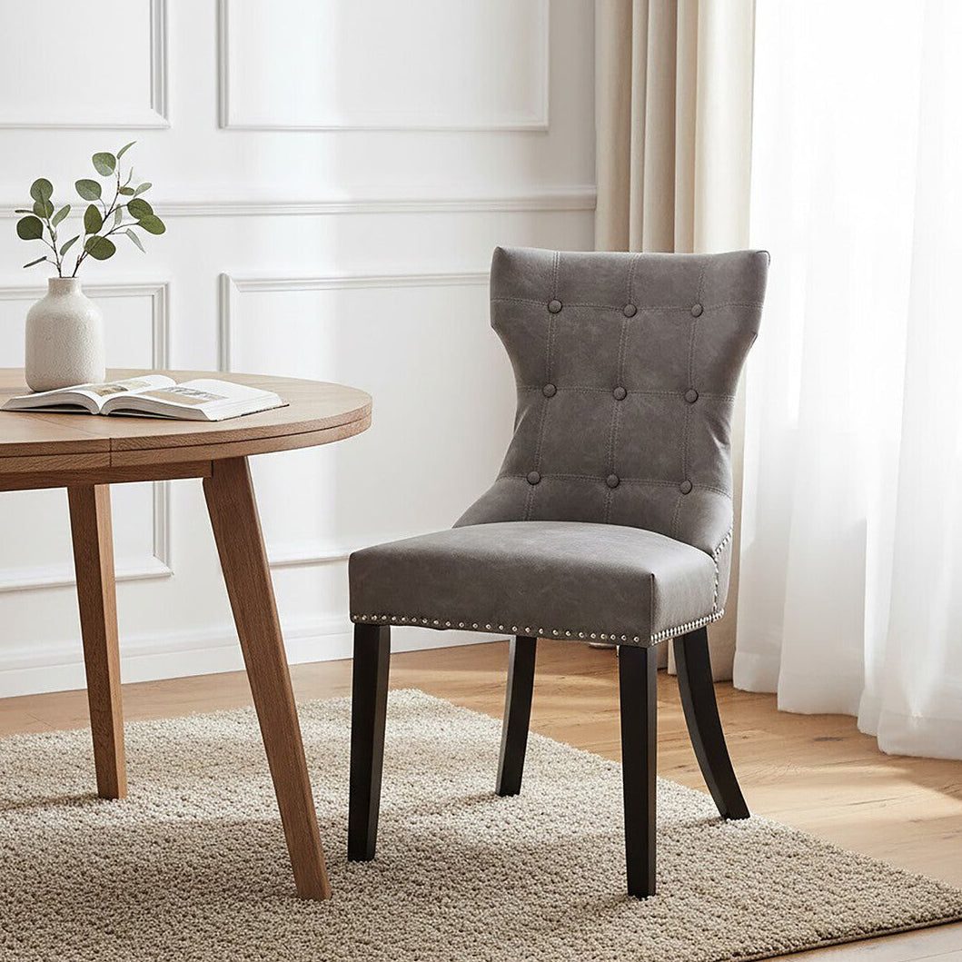 Gray upholstered chair next to a wooden table with a plant and book in a room with white walls and curtains.