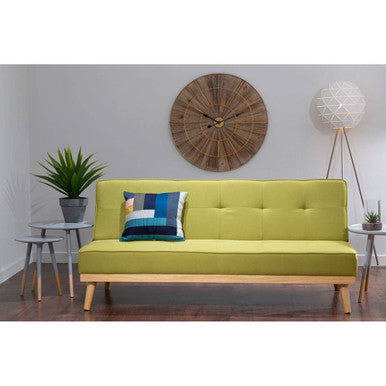 Green sofa with a blue pillow in a room with a clock on the wall.