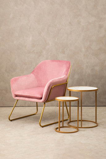 Pink armchair with gold legs and two matching side tables on a beige floor against a neutral wall.