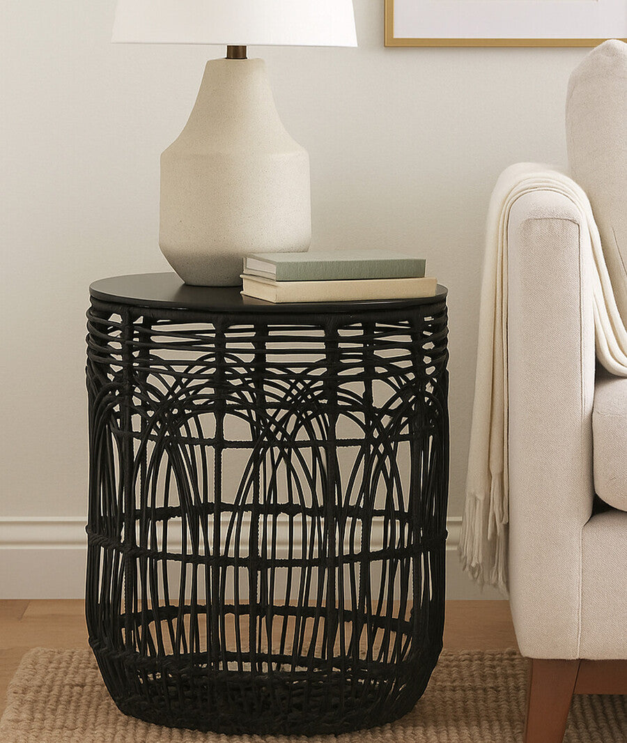 Black woven side table with a lamp and books in a living room setting.