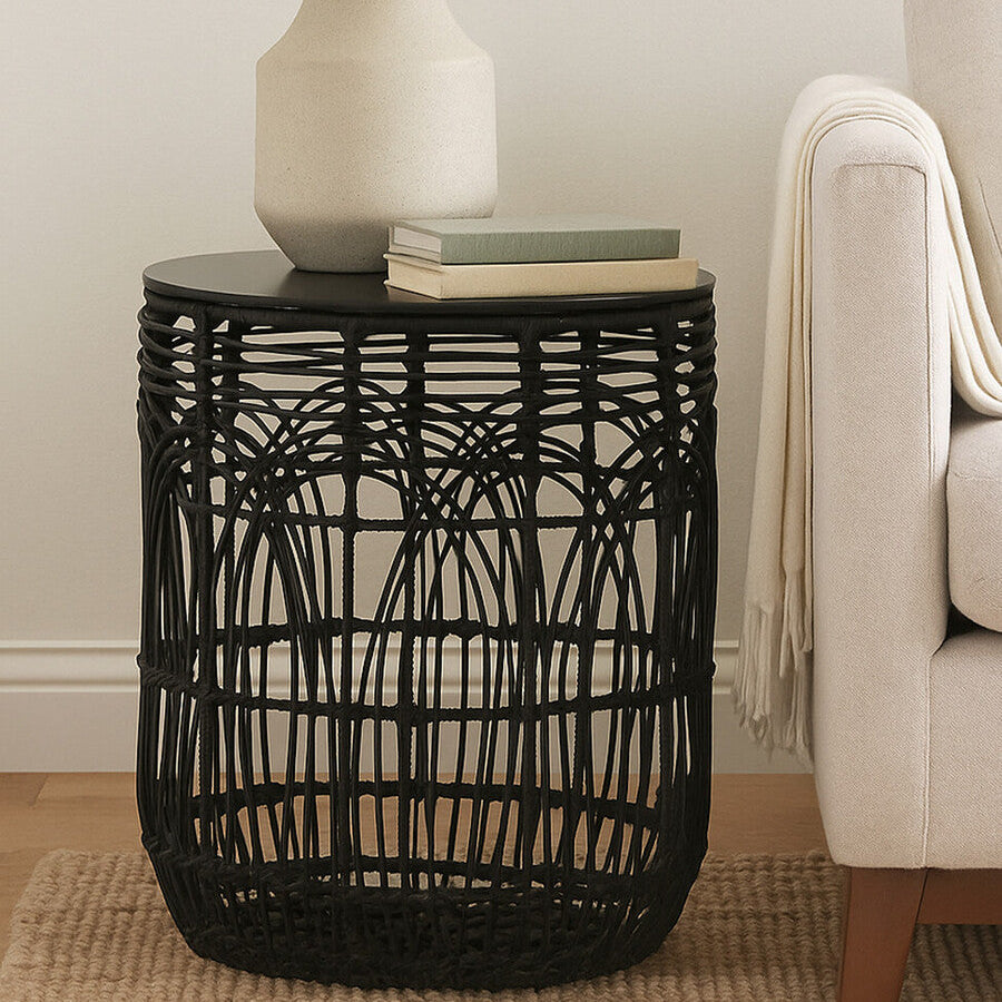 Black woven side table with a lamp and books in a living room setting.