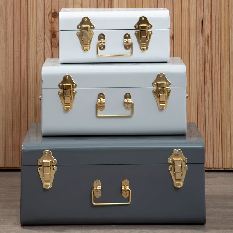 Three stacked trunks in white, gray, and light blue with gold hardware on a wooden floor.