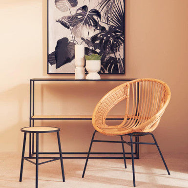 Wicker chair and stool with a black and white plant-themed painting on a beige wall.