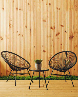 Two black wicker chairs with a small round table on a wooden deck against a wooden wall.