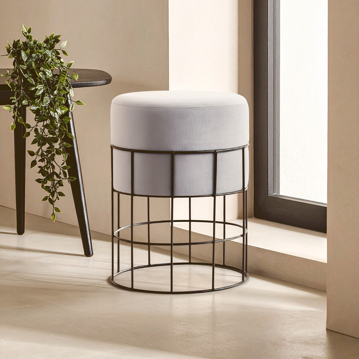 Gray cushioned stool with black metal frame in a room with a plant and table.