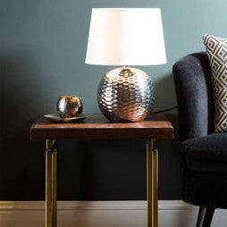 Table lamp with decorative base on a wooden side table against a dark green wall.
