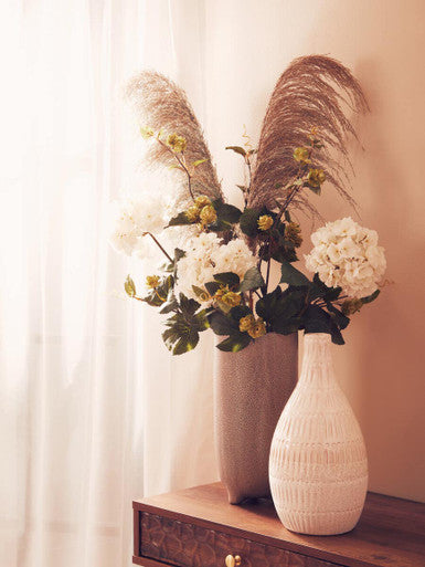 Decorative vase with flowers and pampas grass on a wooden surface with a light curtain background.