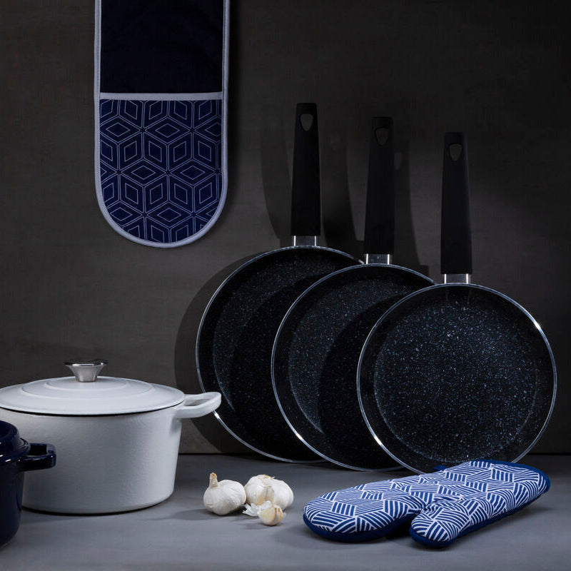 Set of black frying pans with a white pot, garlic, and blue patterned oven mitts on a dark background.