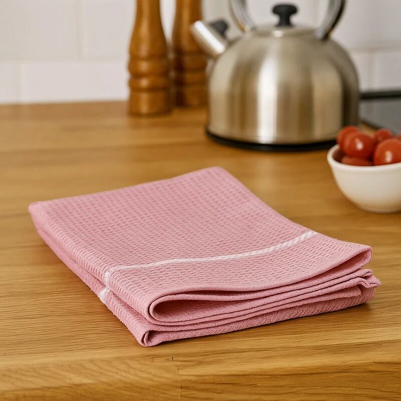 Folded pink towel on a wooden kitchen counter with a kettle and tomatoes in the background.