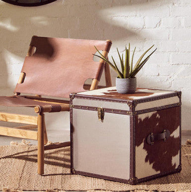 Vintage-style trunk with cowhide accents next to a leather chair against a white brick wall.