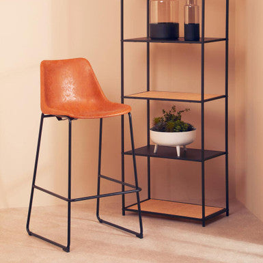 Orange bar stool next to a black metal shelf with decorative items against a beige wall.