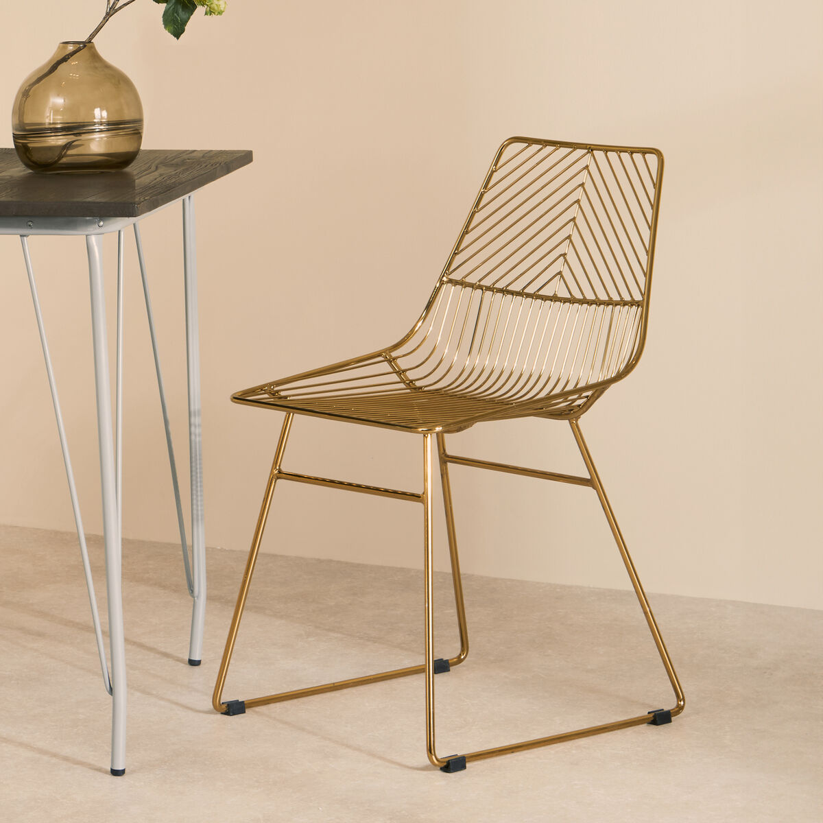 Gold wire chair next to a small table with a vase on a beige background