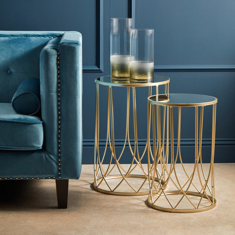 Gold side tables with clear glasses on a blue sofa against a blue wall.