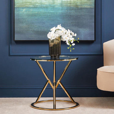 Glass side table with gold legs against a blue wall with abstract painting