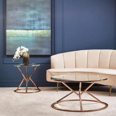 Modern living room with beige sofa, round bronze table, and abstract painting on a blue wall.