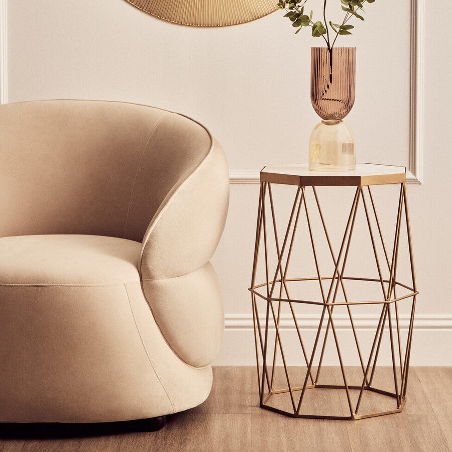 Beige armchair next to a geometric metal side table with a vase on a light-colored wall background