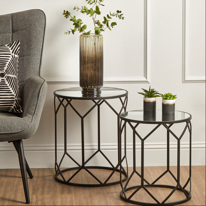 Gray armchair with geometric pillow, two black metal side tables with glass tops, and a vase with greenery.