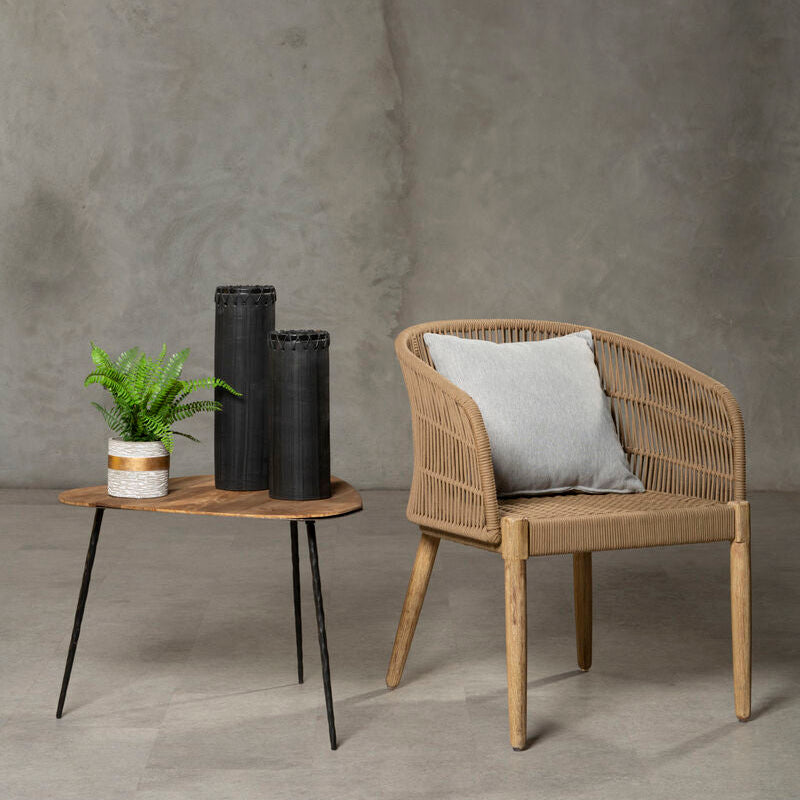 Wicker chair with a cushion and a small round table with a plant against a gray wall.