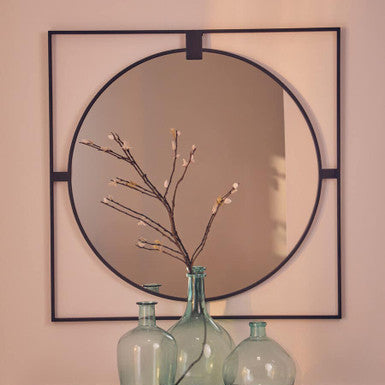 Decorative mirror with a square frame and glass bottles on a table against a beige wall.