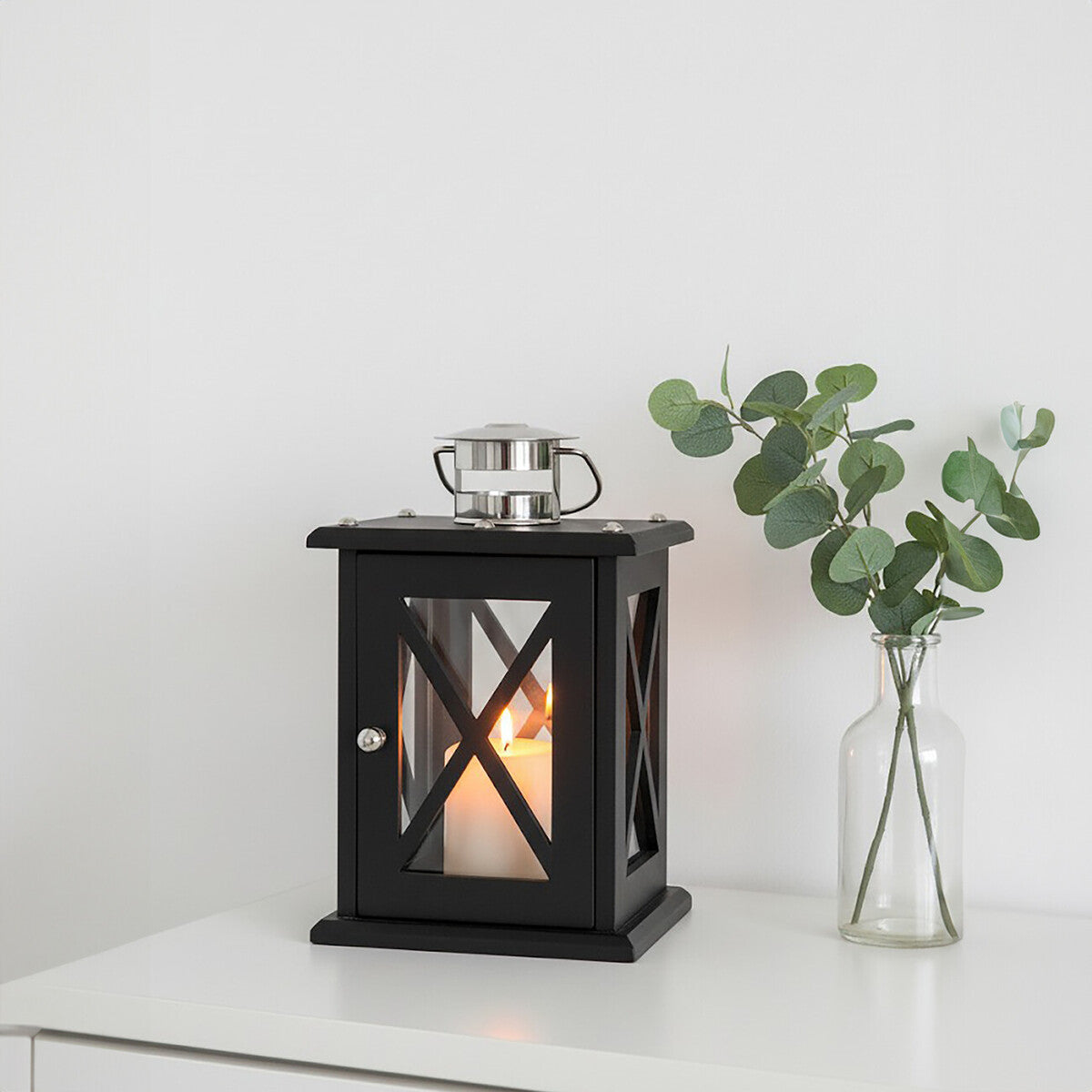 Black lantern with candle on a white surface next to a silver cup and a vase with greenery.