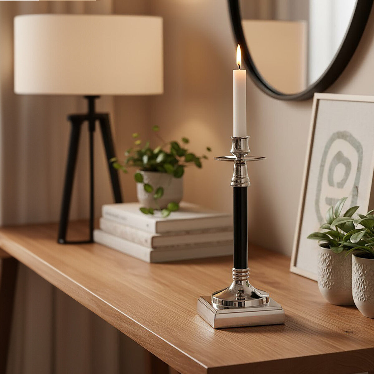 Decorative candlestick with a lit candle on a wooden surface, surrounded by books, plants, and a lamp.