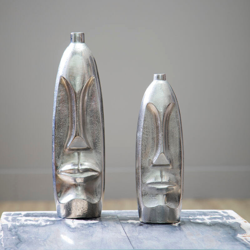 Two metallic face-shaped vases on a reflective surface with a neutral background