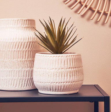Two textured white planters with a small plant on a dark surface against a light pink wall with a round mirror.