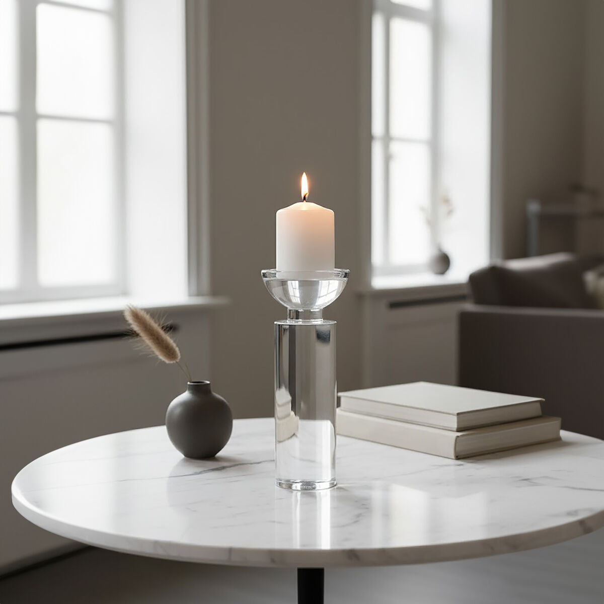 Candle in a glass holder on a marble table with books and a vase in a softly lit room.