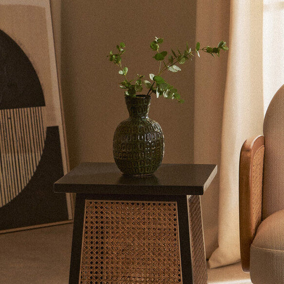 Wicker side table with a textured vase and plant next to a beige armchair in a room with a painting on the wall.