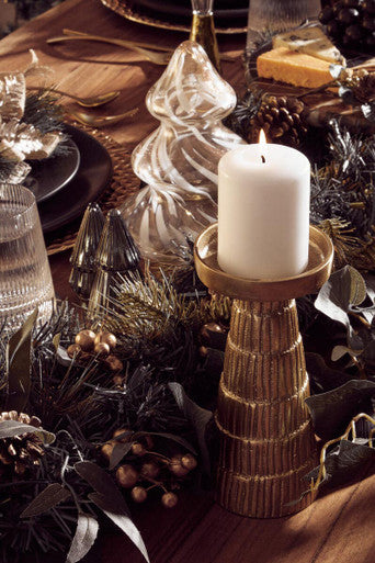 Decorative table setting with a white candle in a gold holder, surrounded by glassware and foliage.